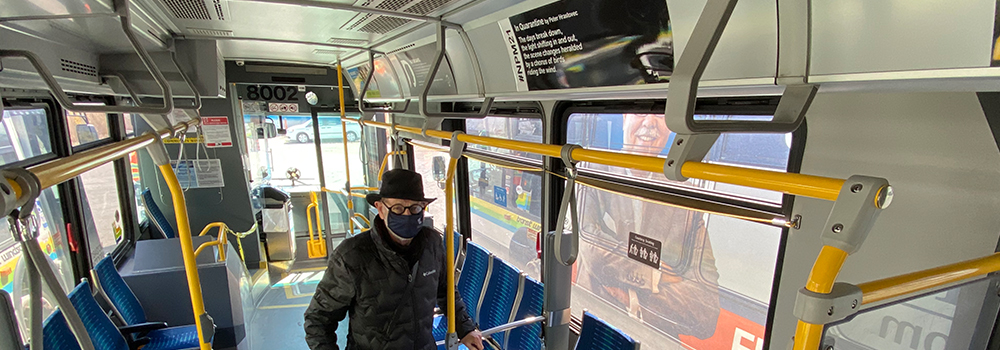 Marty Gervais in a mask, standing under poem on a bus