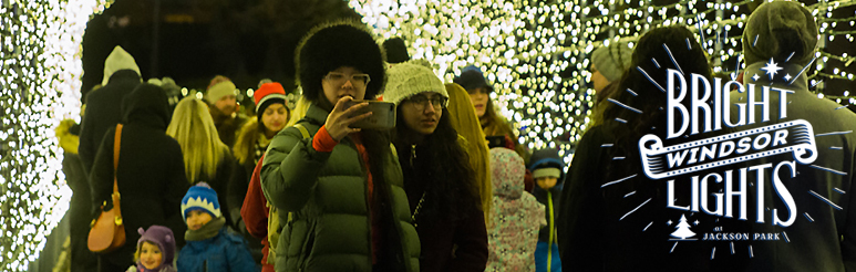 A visitor bundled up for winter taking a picture with a friend in a light tunnel with kids and grown-ups in the background
